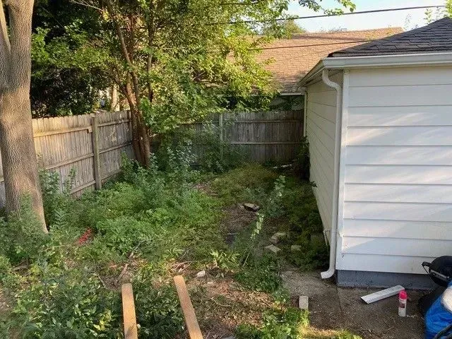 Before photo of a small, overgrown residential backyard area next to a white garage, showing dense weeds and a wooden fence, awaiting a hardscape transformation.