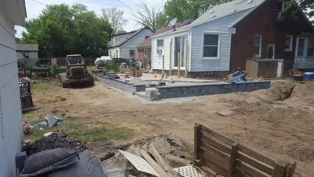 Construction in progress for a residential backyard hardscape patio project. The image shows the outline of a raised patio with retaining walls made of stacked gray blocks, along with construction equipment and dirt.