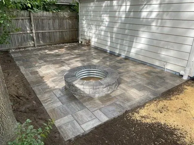 Close-up view of a finished residential paver patio hardscape design, featuring gray and brown pavers surrounding a circular stone fire pit with a metal ring.