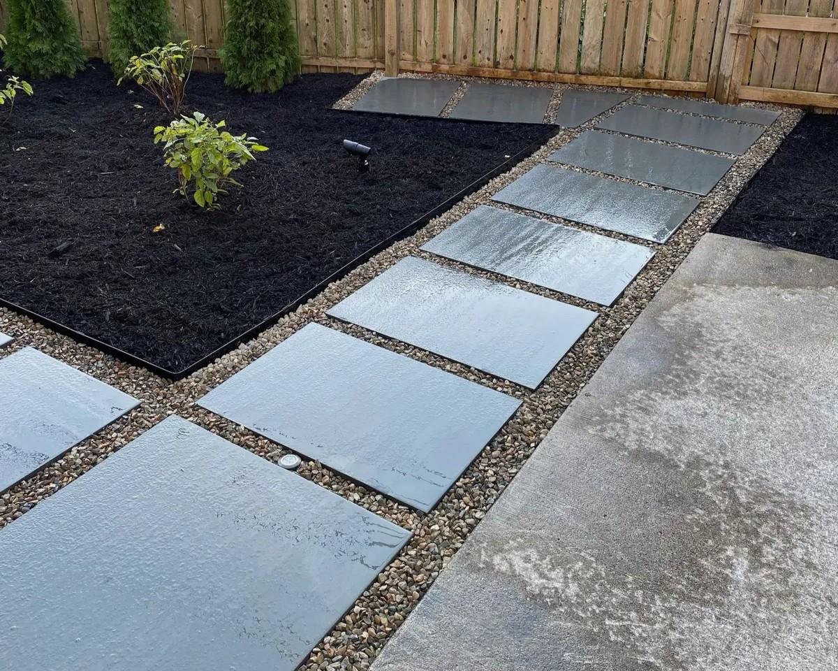 Modern residential landscape design with a stepping stone walkway made of large gray pavers set in river rock, bordering a mulched garden bed.
