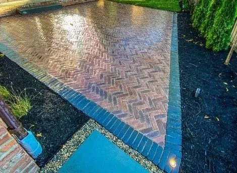 High-gloss residential hardscape patio featuring brick pavers installed in a herringbone pattern with a dark contrasting paver border, next to a sunroom.