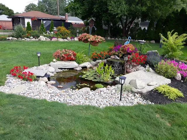 Beautifully landscaped residential water garden featuring a small pond with a waterfall, surrounded by colorful flowers, large stones, white river rock, and solar lights.