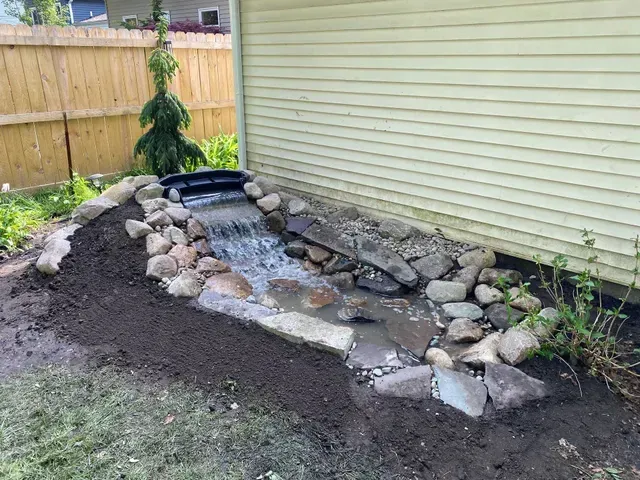 Finished residential backyard water feature in Clinton Township, showcasing a small waterfall and stream cascading over various sized stones and rocks next to a house.