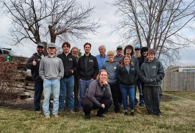 Group photo of a large landscaping and outdoor services team from The Guy Outdoor Services, smiling in front of a yard and trees, used for team recruiting.