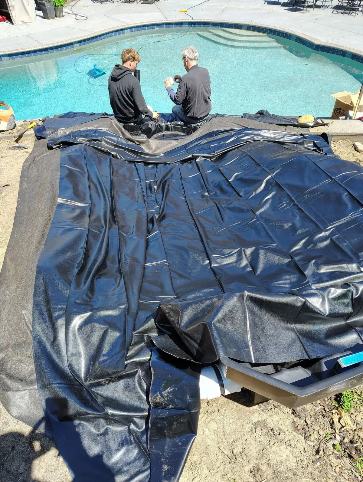Landscaper team members working on a water feature installation next to a swimming pool, showing a large black pond liner being laid out.