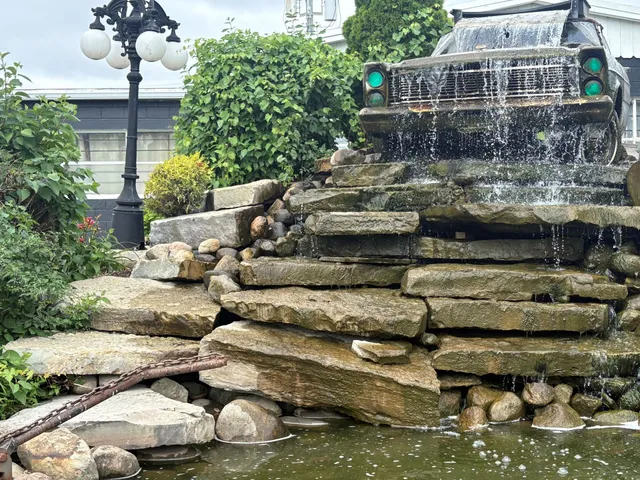 Unique commercial water feature installation featuring a car-themed waterfall where water flows over large stacked flagstones, cascading from the grille of an old car into a pond.
