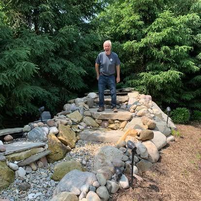 Photo of the business owner standing on top of a newly completed commercial water feature installation, which is a rock waterfall and stream surrounded by large stones and plantings.