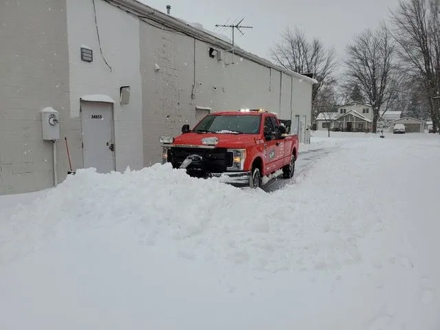 Red snow plowing truck clearing a large amount of deep snow from a commercial business driveway and parking lot during a winter storm.
