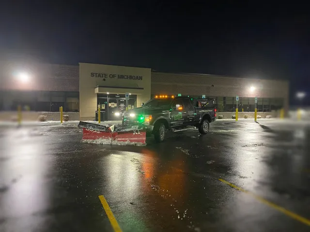 Commercial snow plowing service truck with a plow clearing a large, wet parking lot at the State of Michigan building at night.