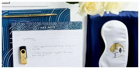 Top view of a desk with a gold USB flash drive, a notepad with notes, a pen, a sleep mask, and white roses.