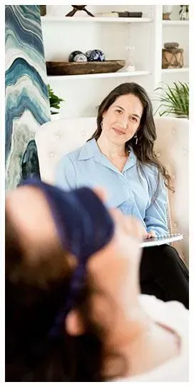 Therapist in light blue blouse taking notes while a blindfolded patient lies on a couch