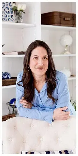 Therapist in light blue blouse leaning on a white chair, smiling in a cozy room with shelves in the background.