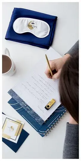 Person writing in a notebook with a gold USB flash drive, sleep mask, and cup of coffee on the desk