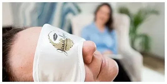 Close-up of a person wearing a white eye mask with Jamie Lynn Hypnotherapy logo, with a therapist in the background.