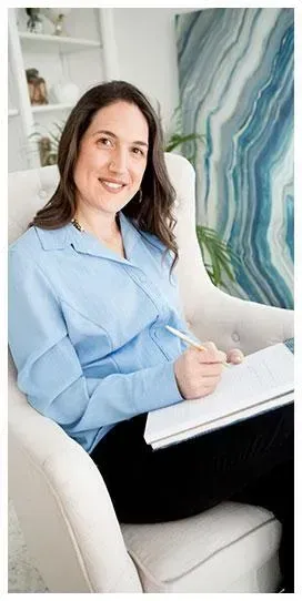 Therapist in light blue blouse sitting in a chair, taking notes with an abstract painting in the background.