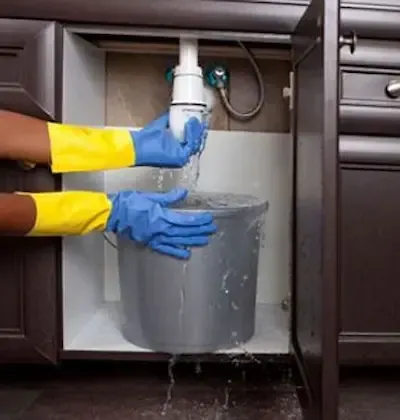 Gloved hands catching water from leaking under-sink pipe into bucket during emergency call