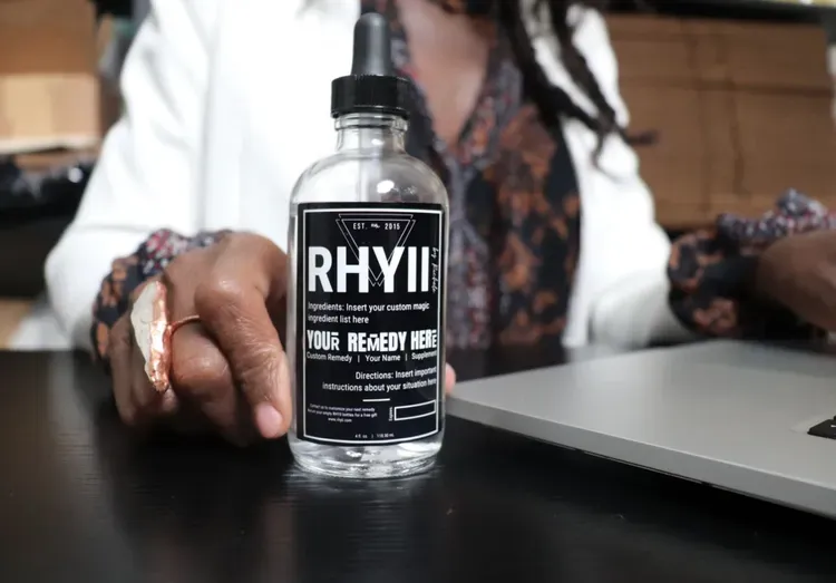 Half portrait photo of Richele Harrison holding a product by RbR Est. 2015 called YOUR REMEDY HERE at a desk for Shoutout SoCal