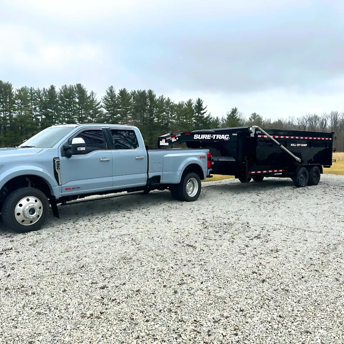 Heavy-duty Ford truck hauling a large Sure-Trac roll-off dumpster on a gravel lot with pine trees in the background.