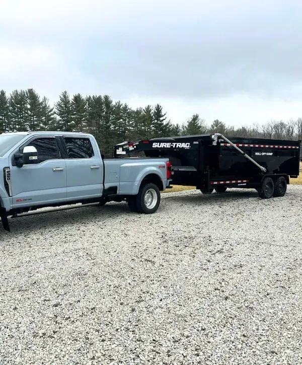 Ford Super Duty truck towing a black roll-off dump trailer on a gravel lot.