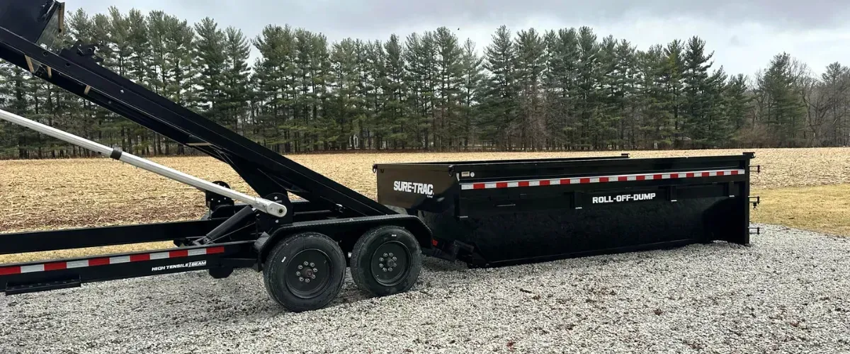 Roll-off trailer dumping a black container on a gravel surface near farmland.