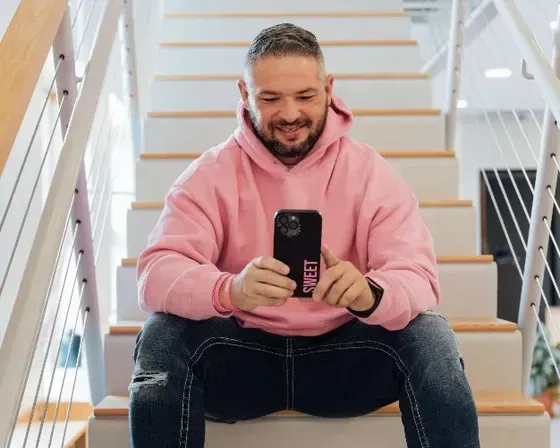 A man wearing a pink hoodie is seated on stairs, focused on his cell phone, conveying a casual and comfortable demeanor