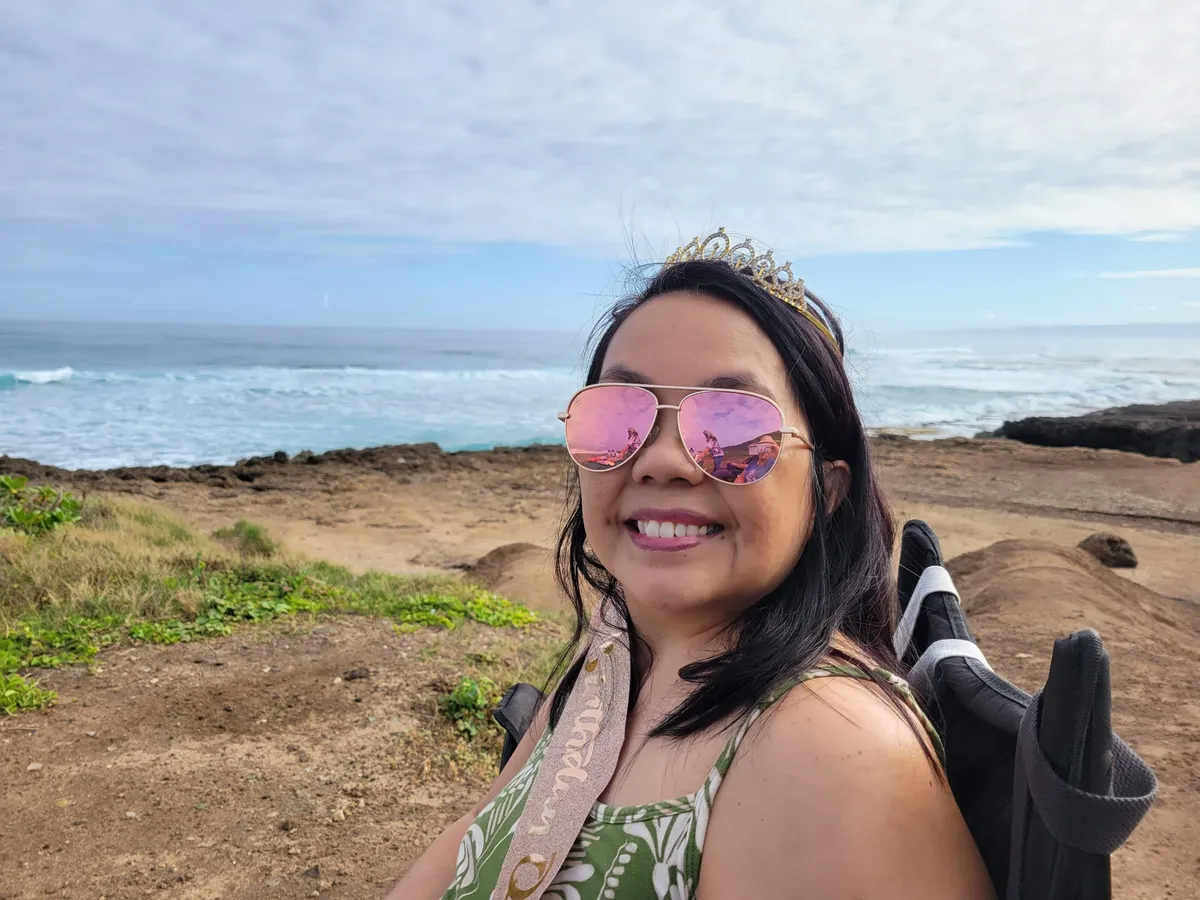 Vince Abramo smiling by the ocean in Hawaiʻi, wearing pink sunglasses and a tiara, enjoying a peaceful coastal moment that reflects her season of gratitude, joy, and being present.