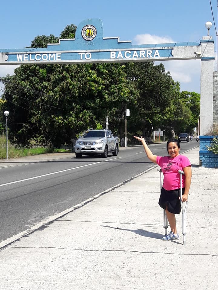 Vince Abramo standing beneath the Welcome to Bacarra archway in the Philippines with her crutches, reflecting on identity, resilience, and living confidently with a limb difference.