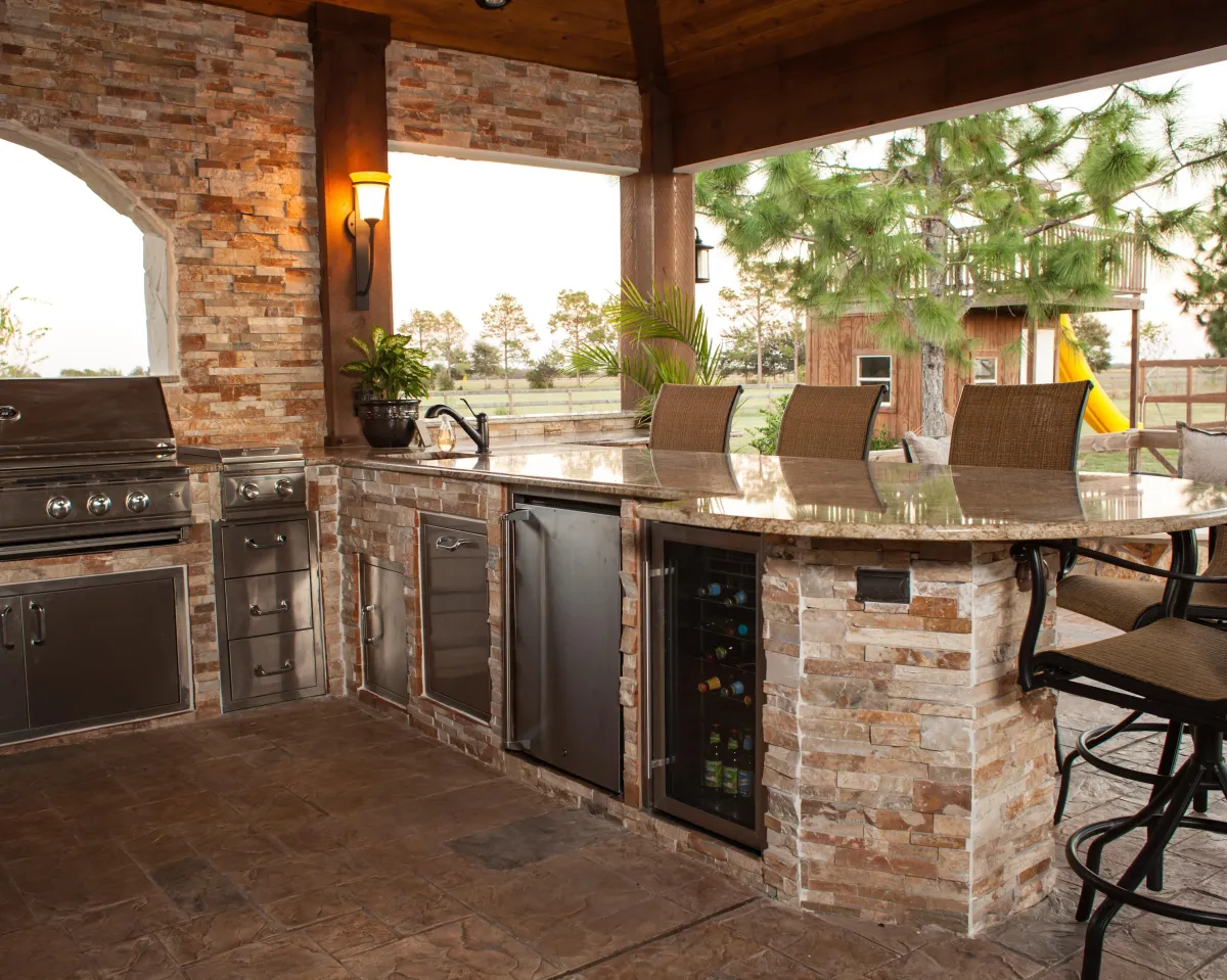 fields by duran: Luxury outdoor kitchen with stone countertops, stainless steel appliances, bar seating, and a view of the backyard and playhouse.
