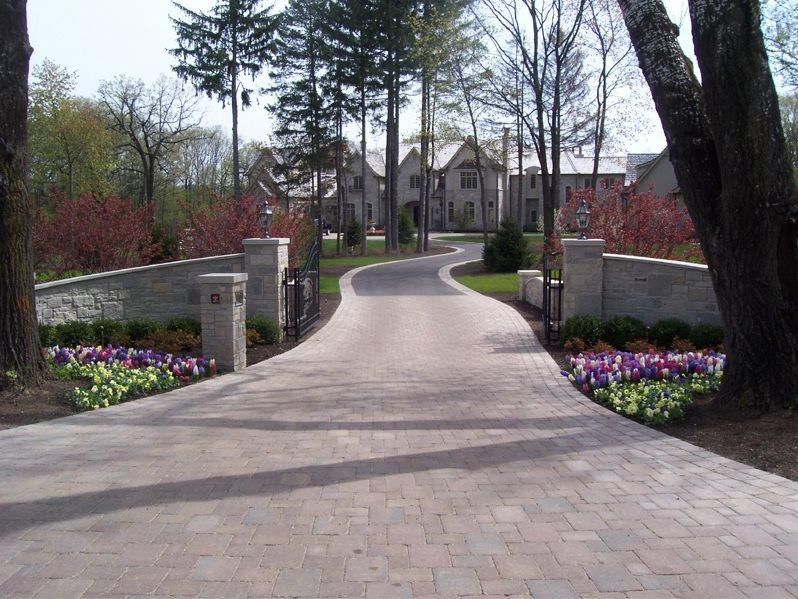 fields by duran: Elegant paver driveway leading to a large estate home, lined with stone walls, flower beds, and mature trees.
