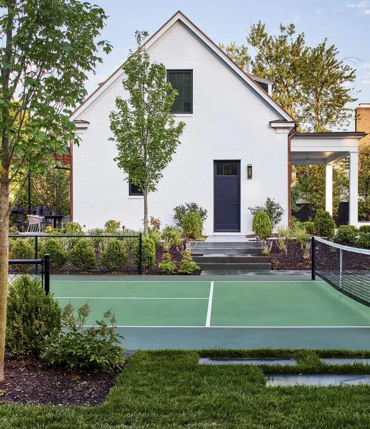 fields by duran: Modern backyard with a small tennis or pickleball court, manicured landscaping, and a white brick house with black door and windows.