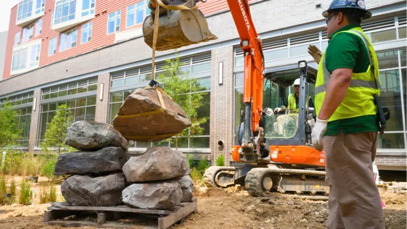 fields by duran: Construction workers using an excavator to place large boulders in a commercial landscape project.