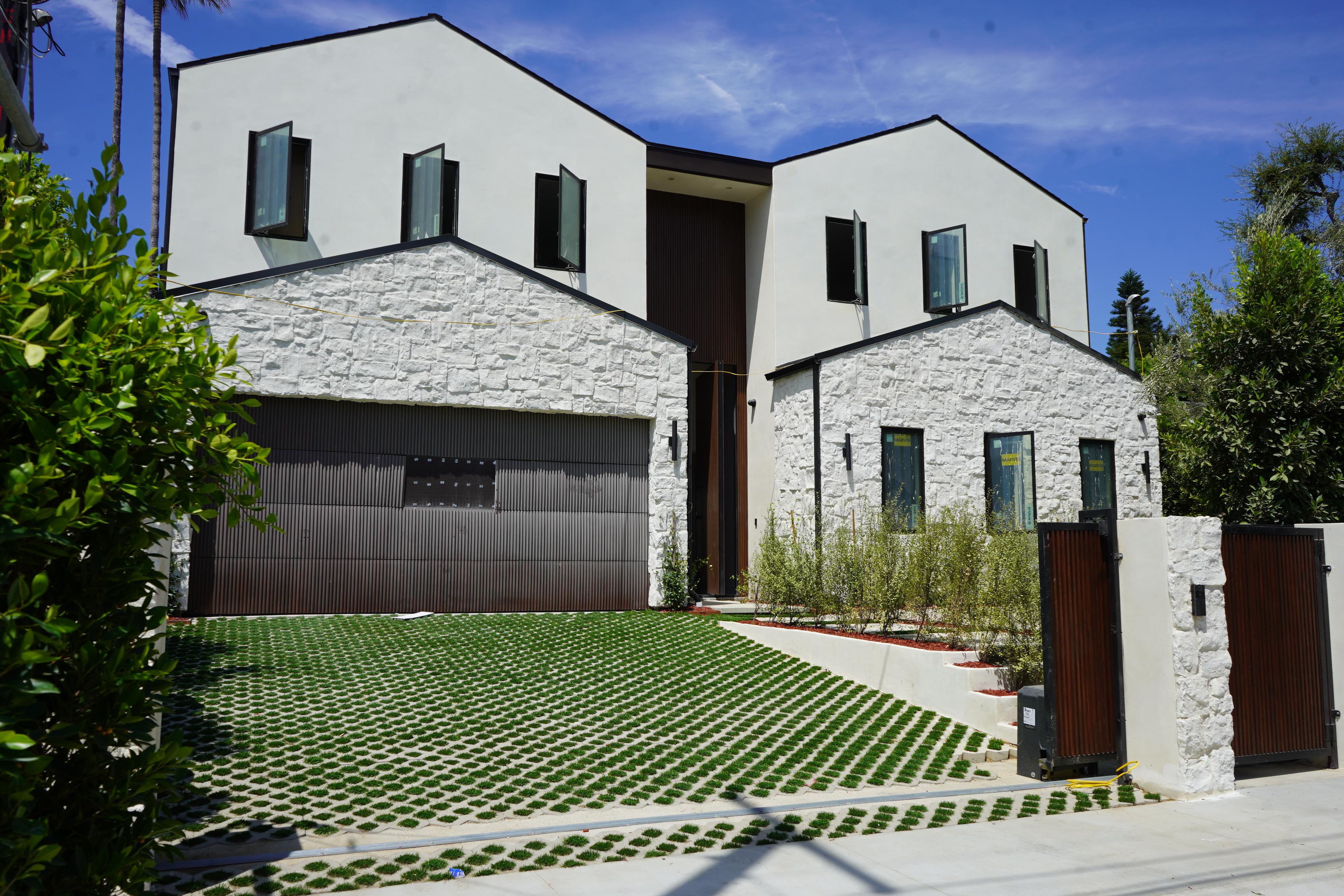 fields by duran: Elegant paver driveway leading to a large estate home, lined with stone walls, flower beds, and mature trees.