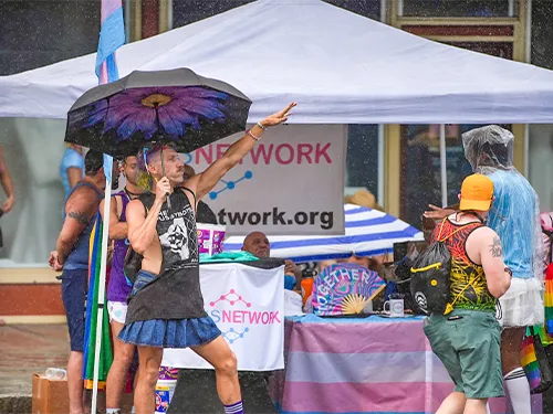 photo of adam morrison outside trasnsnetwork booth at st pete pride 2024 with skirt and umbrella