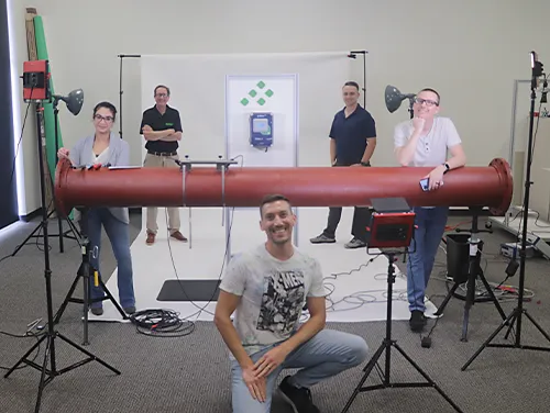 Video shoot at Pulsar Measurement in Largo, Florida, with team members including marketing director Gabriella, senior product manager Ken, and video production specialist (me) in a tie-dye X-Men shirt. The scene features a Pulsar sensor on a large pipe and a branded Pulsar meter on a mock wall, with photography and graphic design setup by Harry Jamison