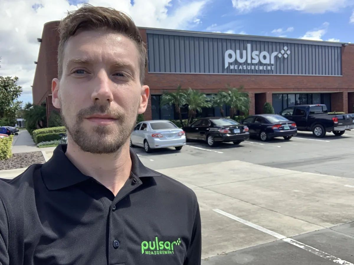 Adam wearing a Pulsar polo in front of Pulsar Measurement's office building with the company logo on the exterior.