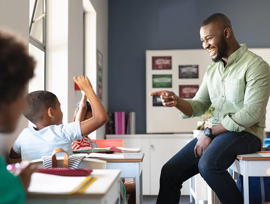 Male teacher in light green shirt engaging with enthusiastic elementary students raising hands in modern classroom