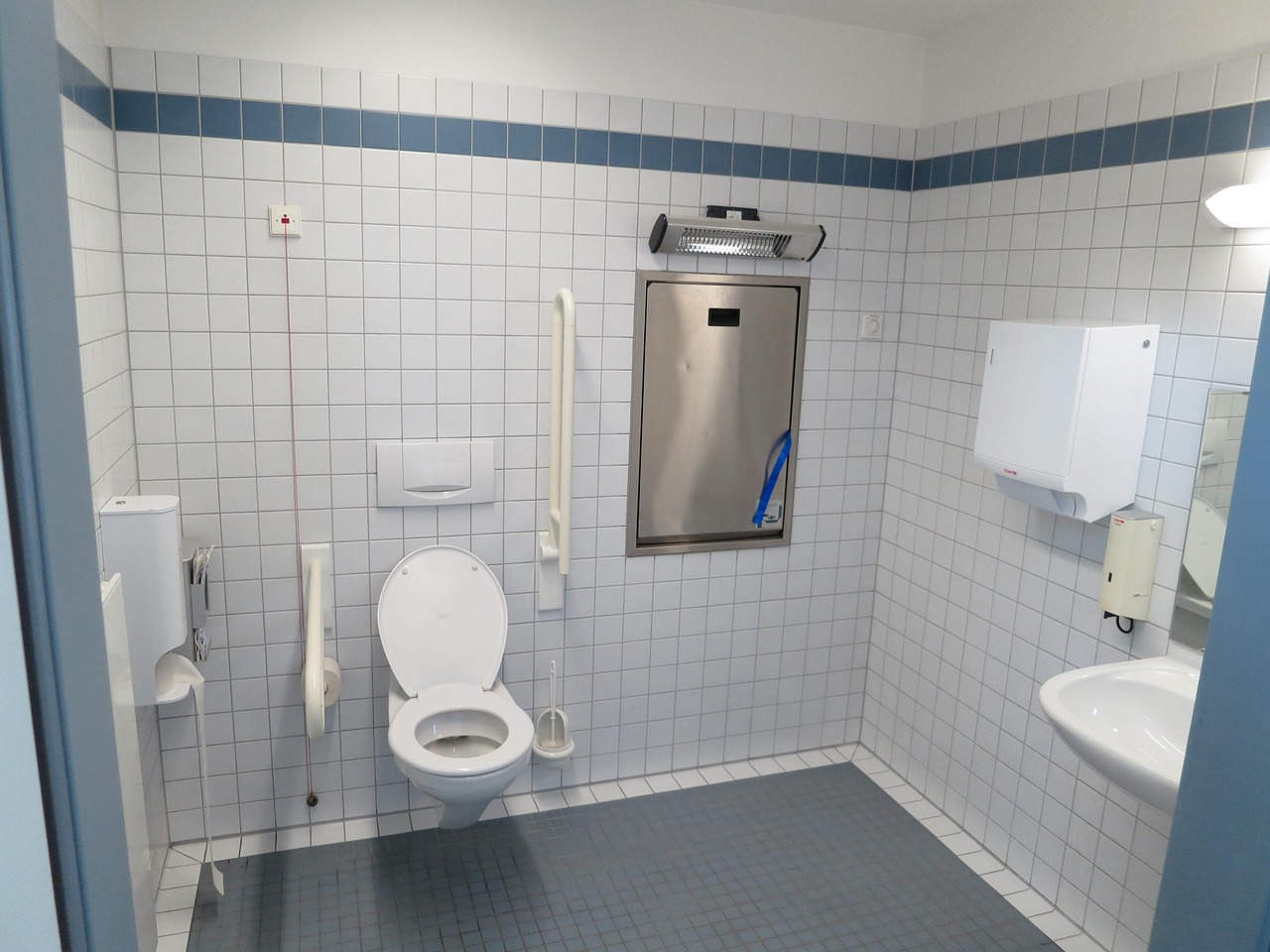 Interior of a commercial bathroom. The space has white tiled walls with a blue stripe, a wall-mounted toilet with a grab bar, a sink, and a metal baby changing station on the wall.
