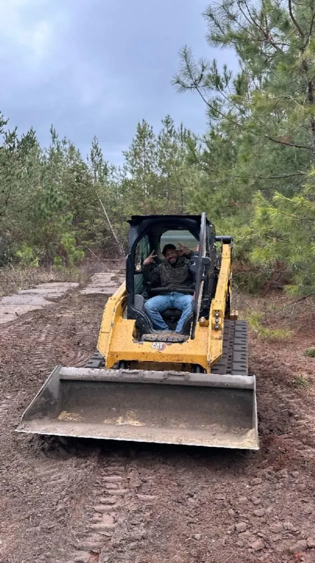 Founder of Vaquero Landworks, Jose Espejo, operating a yellow skid steer loader on a dirt path surrounded by pine trees during a land clearing project by Vaquero Landworks in Dublin, Georgia.