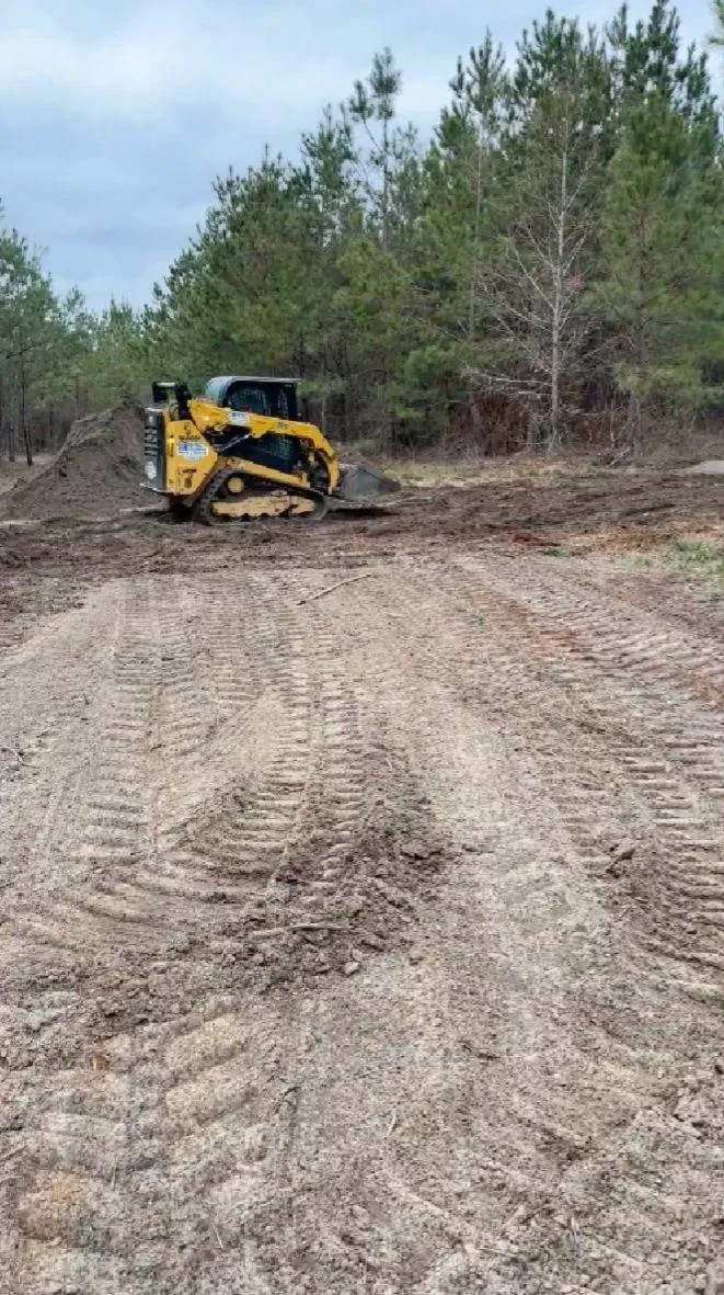 Yellow skid steer grading a dirt path through pine woods, leveling land as part of a site preparation project by Vaquero Landworks in Dublin, Georgia.