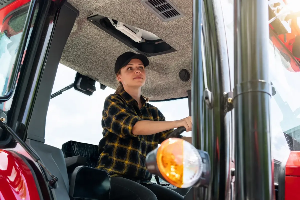 Woman operating a tractor from the driver seat while wearing a black cap and plaid shirt inside farm machinery cab
