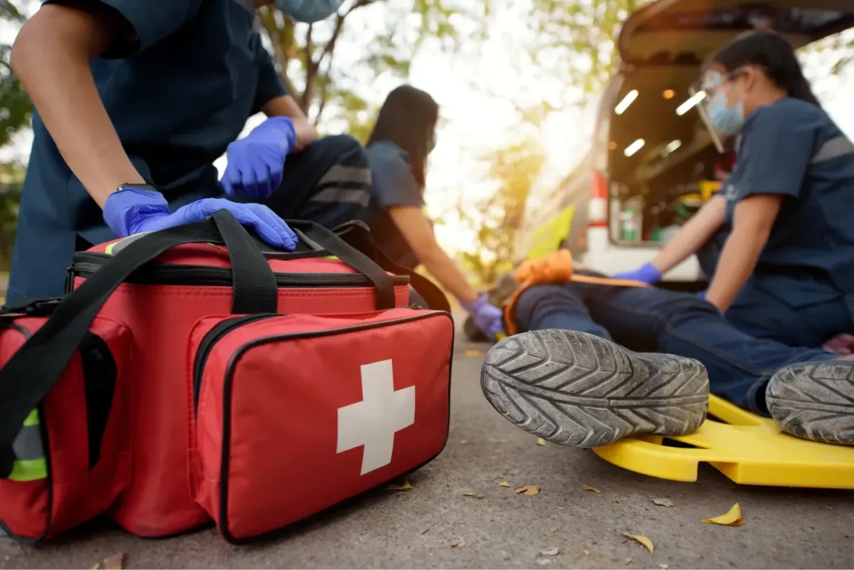 First aid responders assisting an injured person on a stretcher beside a medical kit and ambulance