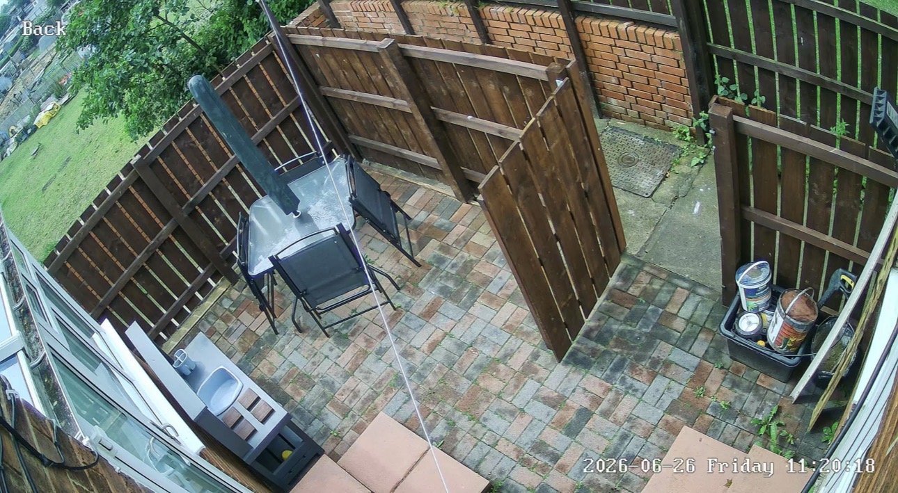 CCTV camera view of a paved back yard with garden furniture and a wooden gate, installed by L&B Security