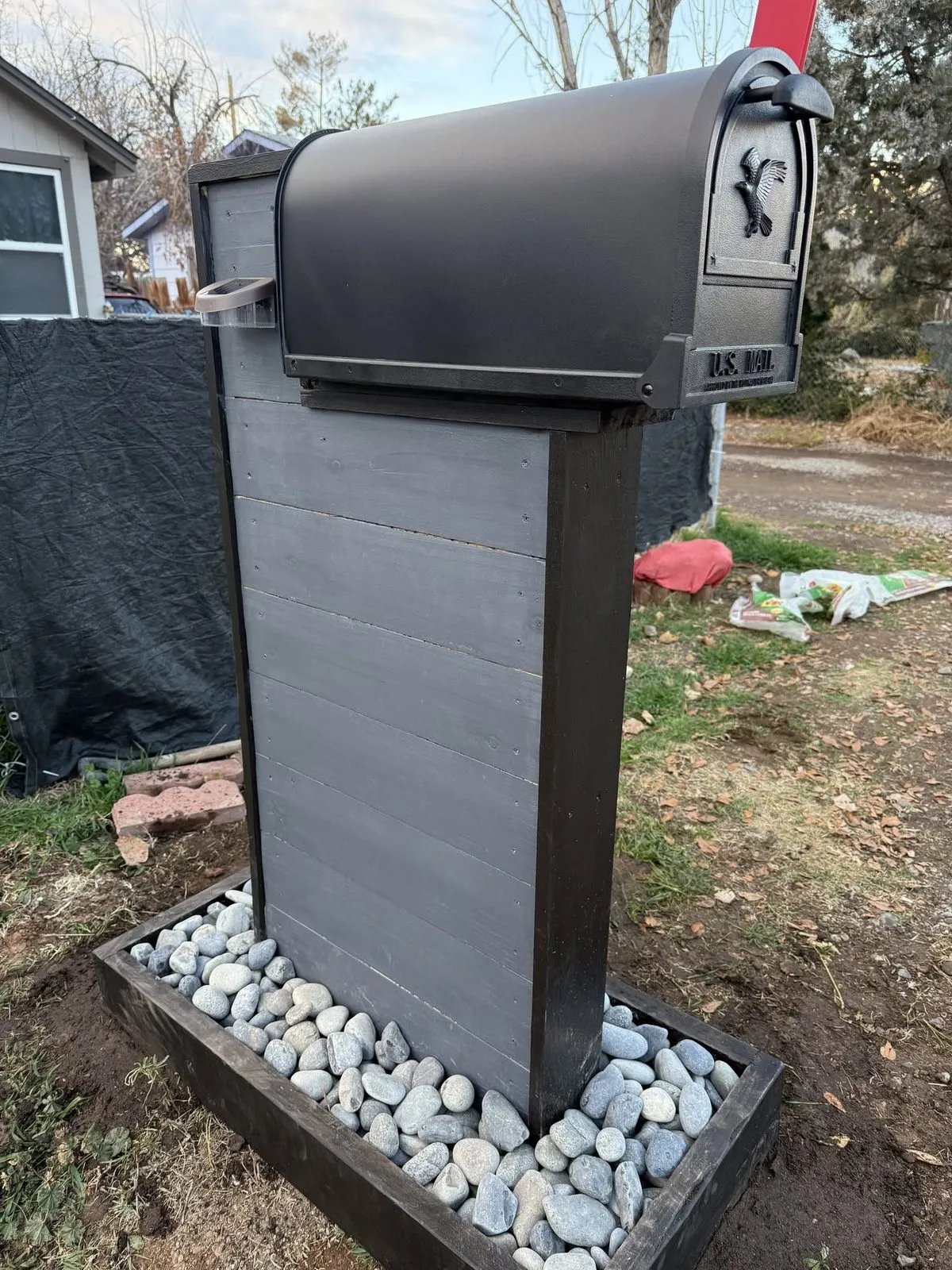 Custom mailbox column installed on a gravel lot with a black mailbox and a river rock base