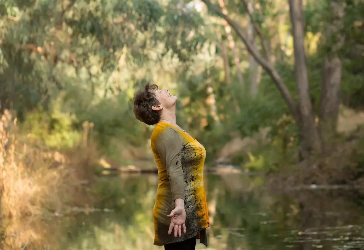 Side view of Suzanne Scurlock standing facing up, palms out, smiling, by a woodland stream.