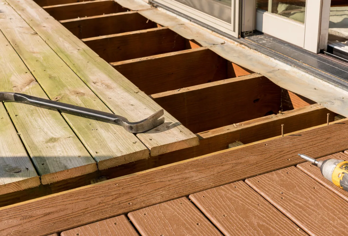 Close-up of a deck repair showing partially removed wooden boards, exposed joists, and tools placed on the surface.