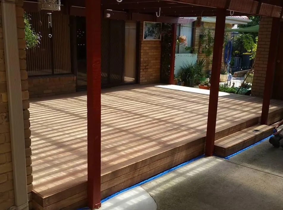 Wooden patio deck built alongside a brick house, partially covered with beams creating striped shadows on the floor.