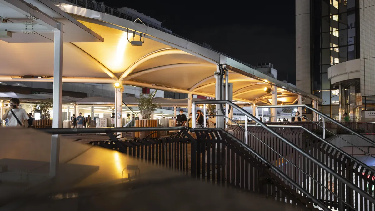 Modern outdoor area at night with illuminated canopy structures, metal railings, and people walking around