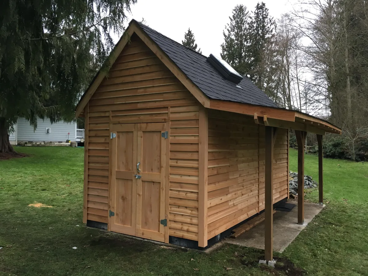 Wooden shed with pitched roof, double doors, and side overhang, located in a grassy backyard surrounded by trees.