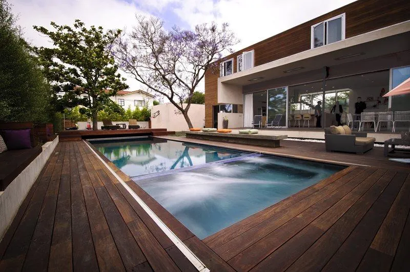 Modern backyard with a rectangular swimming pool surrounded by a dark wooden deck, lounge seating, and a contemporary house