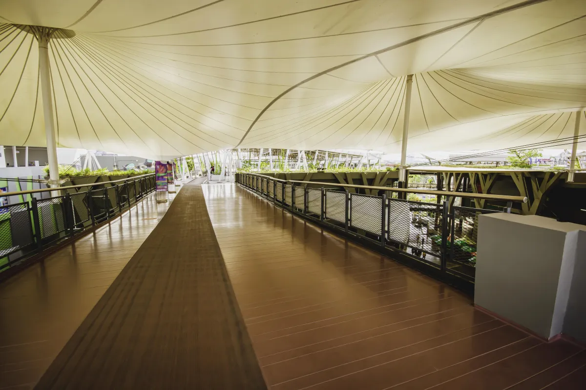 Wide pedestrian walkway with wooden flooring, covered by large white tensile fabric canopies and bordered by railings.